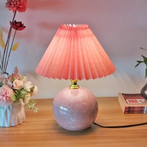Sur une table en bois on voit une lampe en céramique rose avec un abat jour plissé rose. À gauche, il y a un pot blanc avec des fleurs dedans.