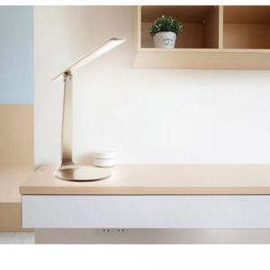 SUr un bureau en bois clair, on voit une lampe de bureau dorée allumée. en haut à droite de l'image, il y a une étagère à case avec une petite plante verte dans un pot blanc.