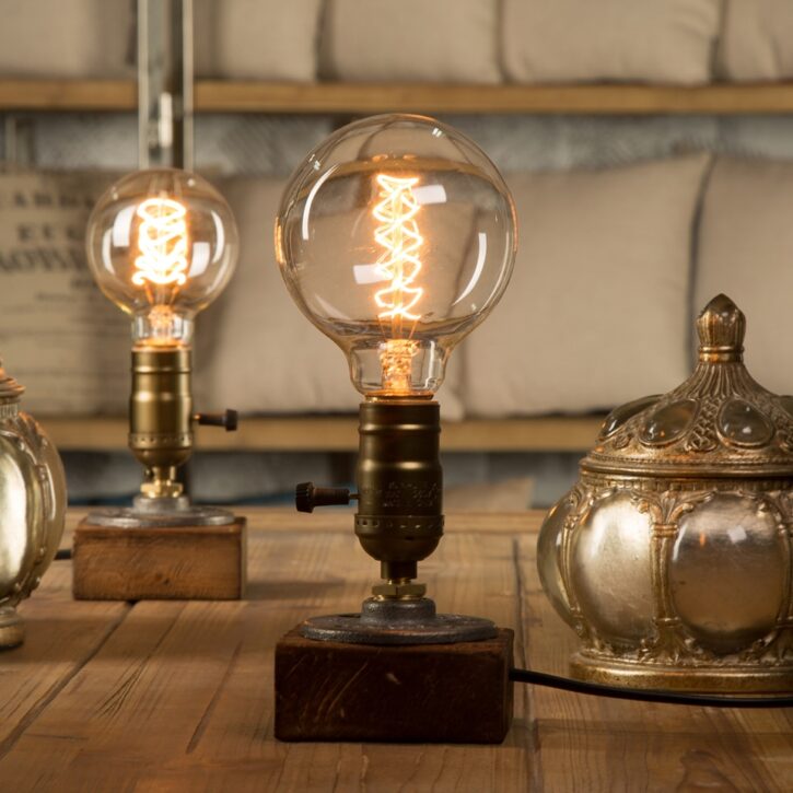 Sur une table en bois on voit au premier plan une lampe industrielle vintage en bois avec un grosse ampoule allumée. Il y en a une autre au second plan à gauche et au second plan à droite une boîte en métal.