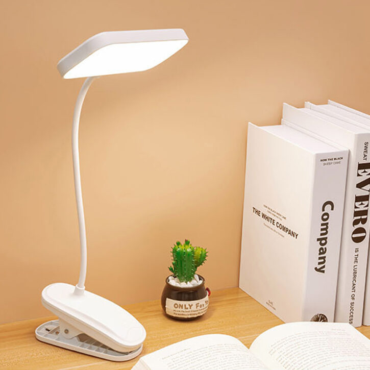 Une lampe blanche à pince posé sur un bureau en bois, devant la lampe un cahier ouvert