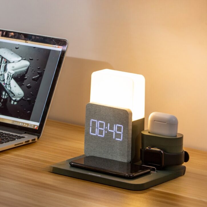 Une lampe réveil posé sur un bureau en bois, un ordinateur portable posé à gauche.