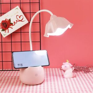 Lampe de bureau rose en forme de fleur avec un support de téléphone et un téléphone posé dessus. La lampe est posée sur un bureau à carreaux rose .