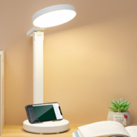 Sur un fond marron on voit une lampe de bureau blache posée sur une table avec un téléphone portable, un petit pot de fleur vert et un livre ouvert
