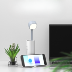 Sur un fond blanc on voit à gauche une petite plante verte, au milieu une lampe blanche avec un téléphone portable et à gauche un morceau d'écran d'ordinateur noir.