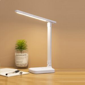 Sur cette image on voit une lampe blanche posé sur une table en bois ainsi qu'une petite plante verte, une feuille blanche et un stylo.