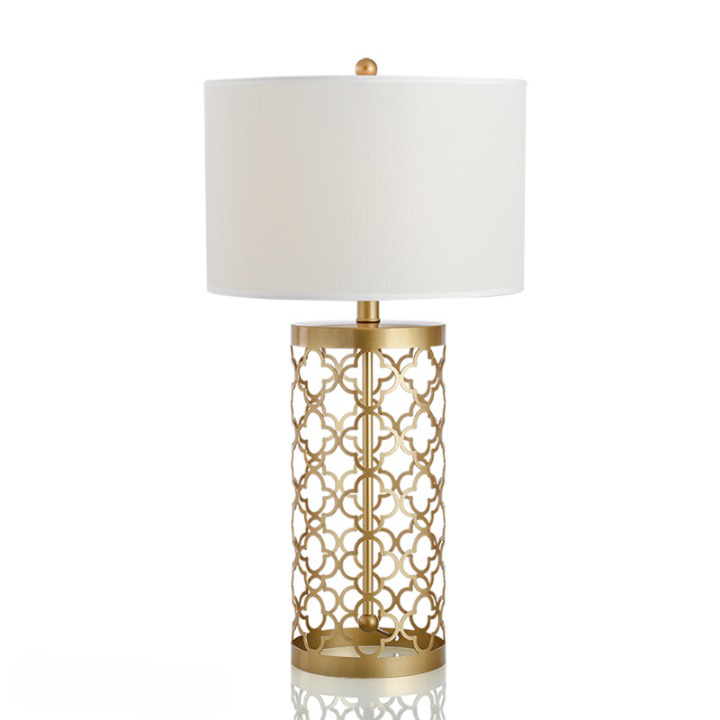 Sur un fond blanc on voit une lampe de bureau avec un abat-jour blanc et un magnifique pied forgé en métal doré.