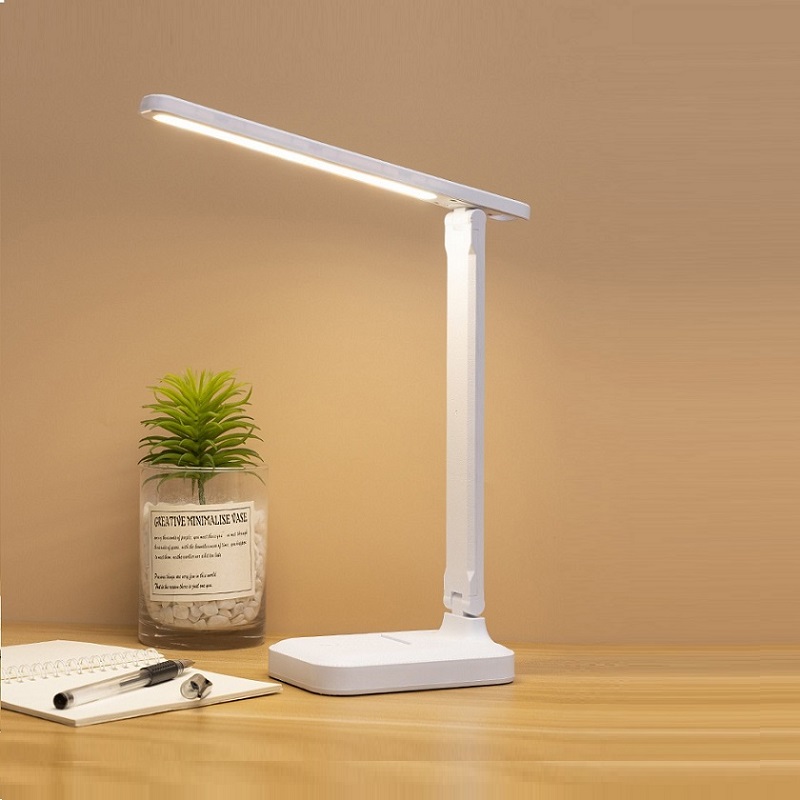 Sur cette image on voit une lampe blanche posé sur une table en bois ainsi qu'une petite plante verte, une feuille blanche et un stylo.