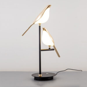 Sur un fond blanc on voit une lampe en forme de deux oiseaux dorés allumée. Le pied de la lampe est noir.