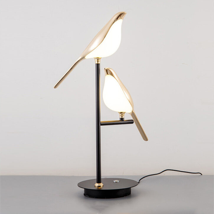 Sur un fond blanc on voit une lampe en forme de deux oiseaux dorés allumée. Le pied de la lampe est noir.