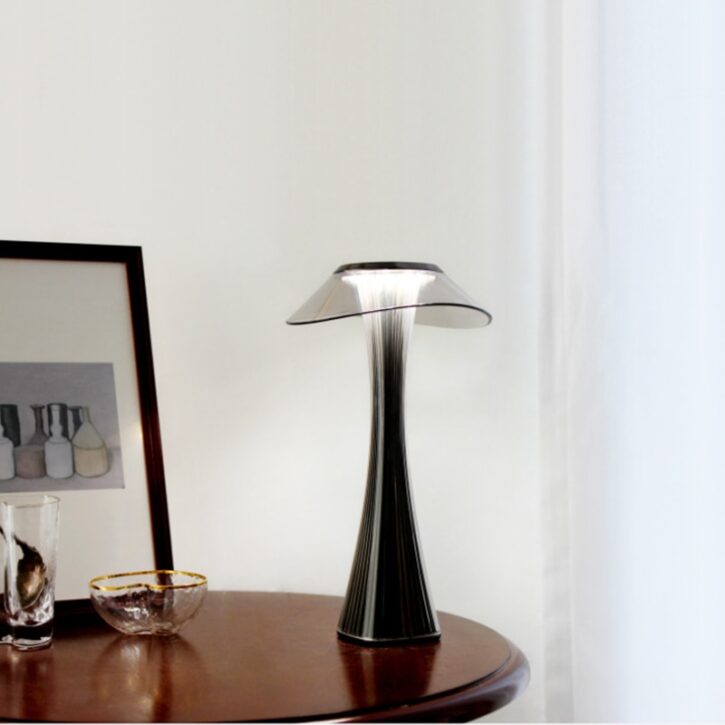 Sur un fond blanc on voit une table en bois marron avec dessus une lampe noire style art-déco et un cadre noir à gauche avec une image dedans. Et sous le cadre un bol transparent.