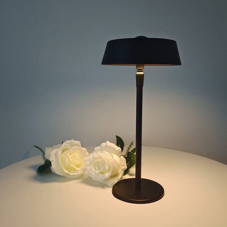 Sur un fond noir on voit une table ronde blanche avec posé dessus une lampe de bureau noire et au pieds de la lampe de roses blanches.