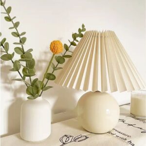 Sur un morceau de table blanche on voit une lampe banche avec un un abat-jour plissé. Et à gauche un vase blanc avec du feuillage vert dedans.