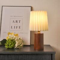 Sur une table on voit une lampe avec le pieds en bois et abat-jour plissé blanc à droite. À gauche on voit un cadre et des fleurs posées devant.